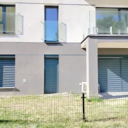 Nowoczesny budynek z szarymi elewacjami i oknami zabezpieczonymi szarymi, zewnętrznymi systemami osłonowymi, widoczne szklane balustrady balkonów i ogrodzenie siatkowe przed budynkiem.