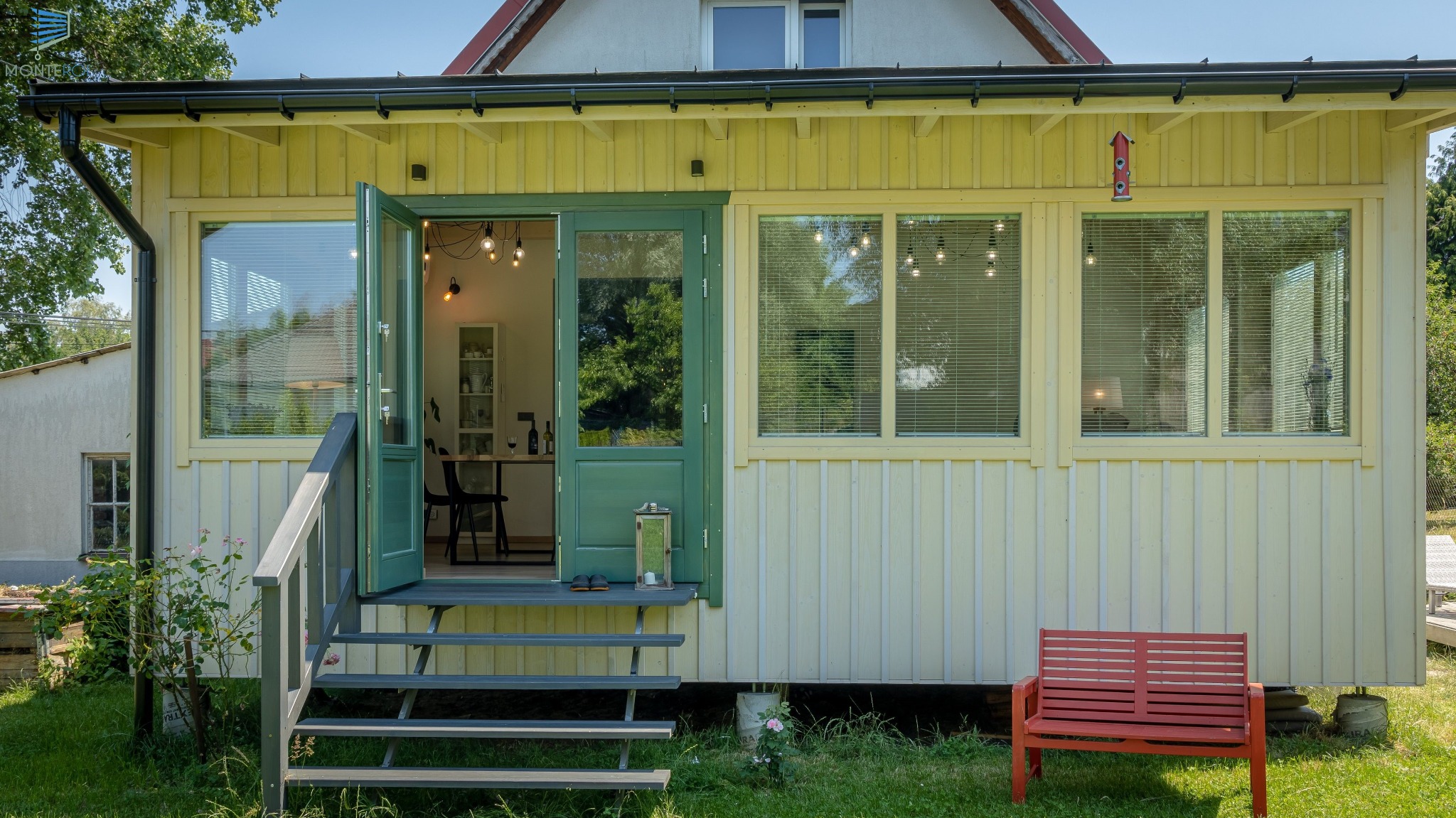 Jasnożółty domek letniskowy z zielonymi drzwiami wejściowymi i oknami z poziomymi żaluzjami, widok z zewnątrz na schody i małą ławeczkę.