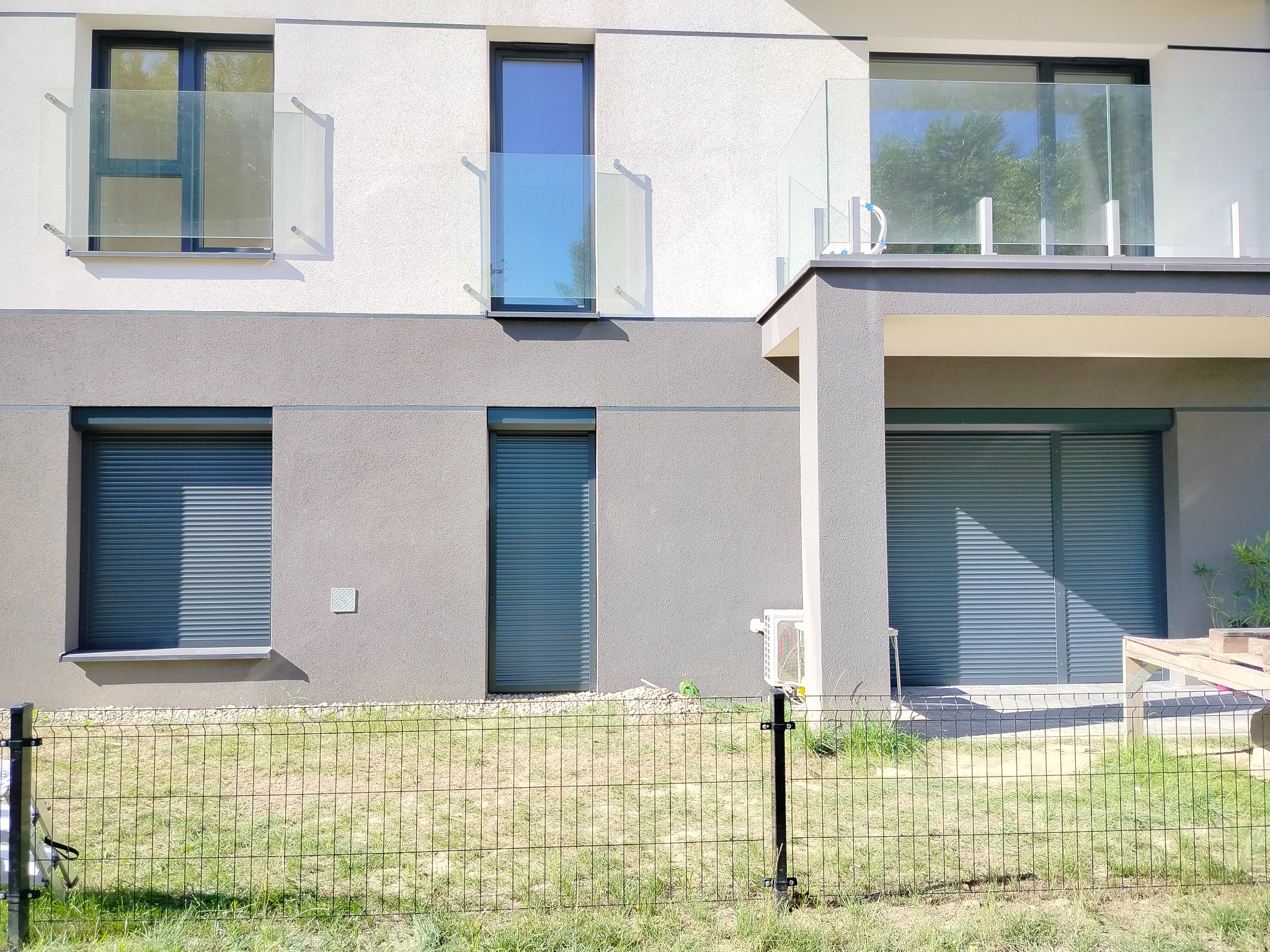 Nowoczesny budynek z szarymi elewacjami i oknami zabezpieczonymi szarymi, zewnętrznymi systemami osłonowymi, widoczne szklane balustrady balkonów i ogrodzenie siatkowe przed budynkiem.