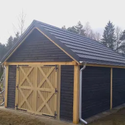 Gotowa wiata z blachodachówką modułową i orynnowaniem metalowym, elewacja z deski świerkowej, dwuskrzydłowe drewniane wrota z czarnymi zawiasami.