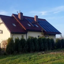 Dom jednorodzinny z panelami słonecznymi na dachu z czerwonej dachówki, drabina oparta o dach, widok z poziomu trawnika.