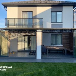 Top Technik sp. z o.o. - Nowoczesny taras z przeszkleniami przesuwnymi, widok z zewnątrz na dwukondygnacyjny dom z balkonem i oknami, na tarasie drewniany stół z ławkami, osoba siedząca przy stole, na trawniku logo firmy.