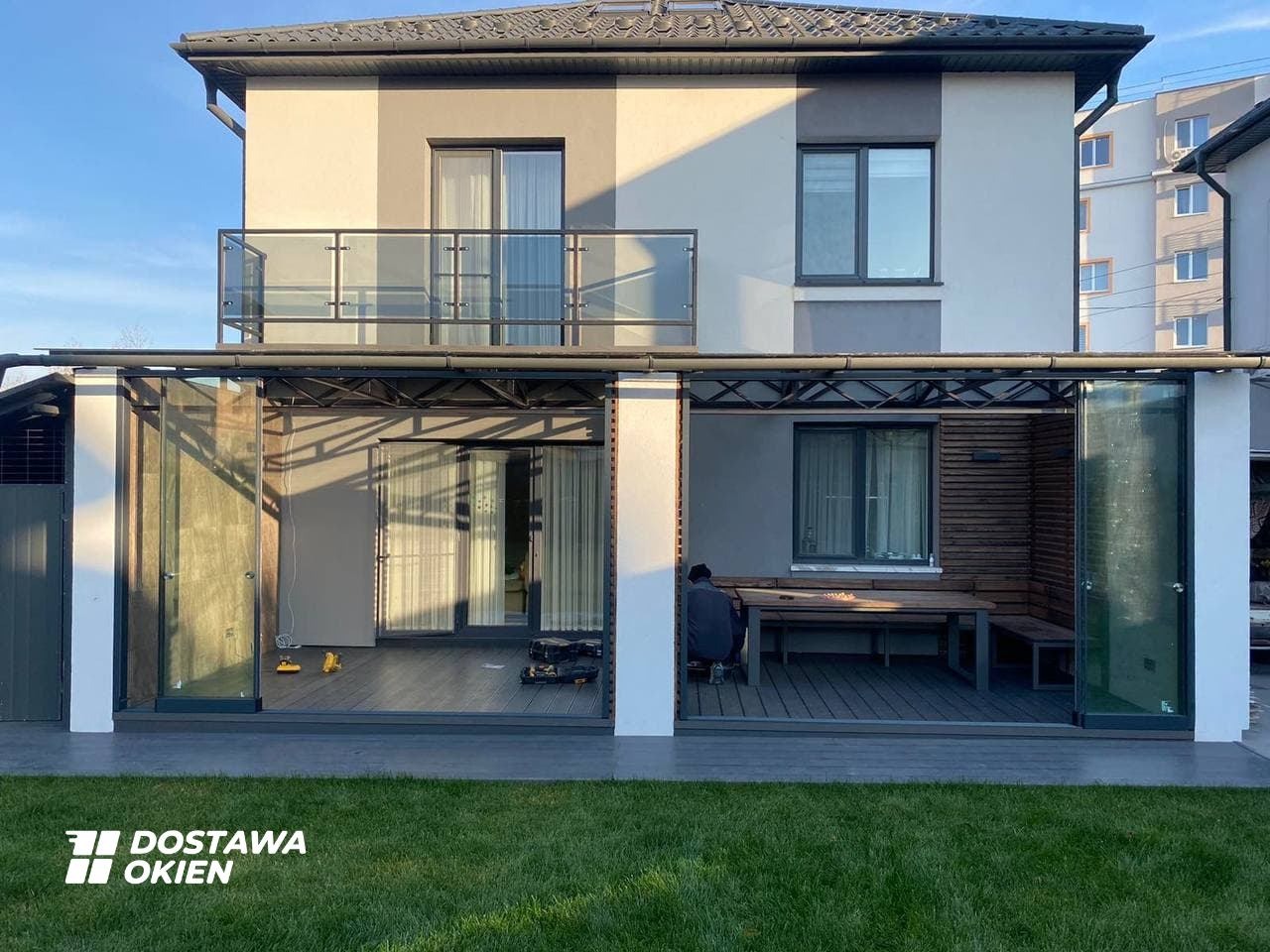 Nowoczesny taras z przeszkleniami przesuwnymi, widok z zewnątrz na dwukondygnacyjny dom z balkonem i oknami, na tarasie drewniany stół z ławkami, osoba siedząca przy stole, na trawniku logo firmy.