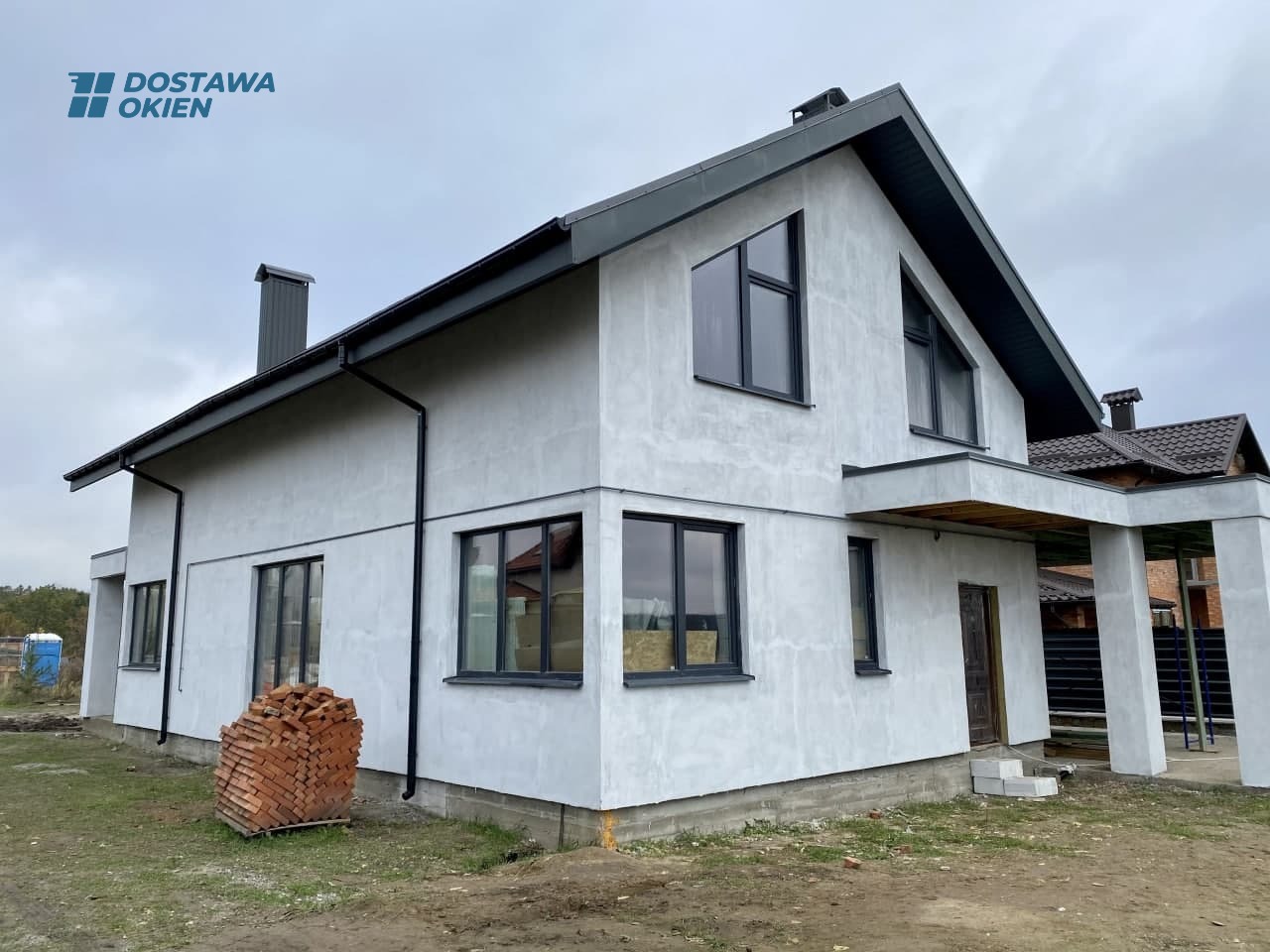 Surowy, dwukondygnacyjny dom w trakcie wykończenia elewacji, z zamontowanymi ciemnymi oknami i stosem cegieł na trawniku.