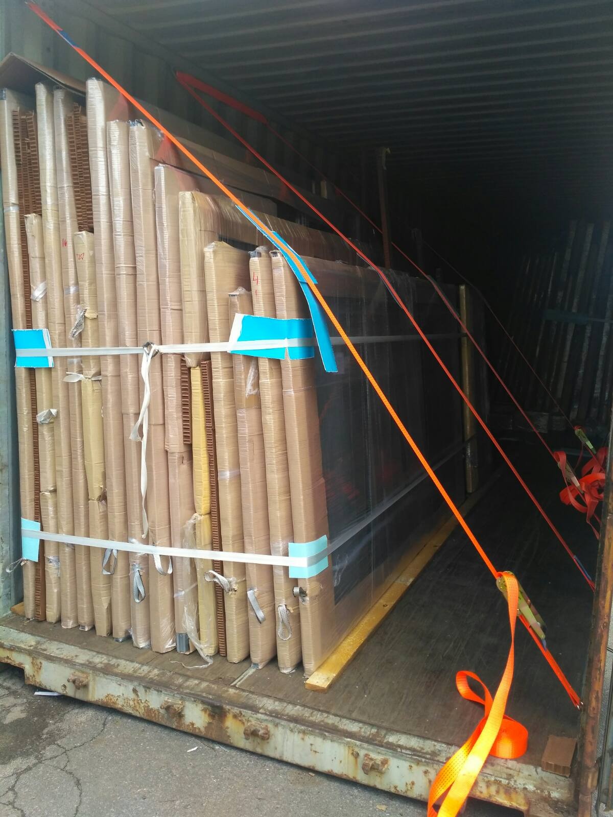 Paczki zapakowanych okien i drzwi zabezpieczone pasami transportowymi w kontenerze, widoczne folie ochronne i kartonowe zabezpieczenia.