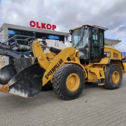 Olkop - Żółta ładowarka kołowa Caterpillar 330D, zaparkowana na kostce brukowej przed budynkiem z napisem OLKOP, z fragmentem frezarki do asfaltu zamontowanej z przodu.