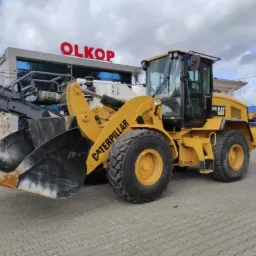 Żółta ładowarka kołowa Caterpillar 330D, zaparkowana na kostce brukowej przed budynkiem z napisem OLKOP, z fragmentem frezarki do asfaltu zamontowanej z przodu.