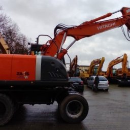 Olkop - Pomarańczowo-szara koparka kołowa Hitachi Zaxis 140W na placu z innymi maszynami budowlanymi, widoczne samochody osobowe