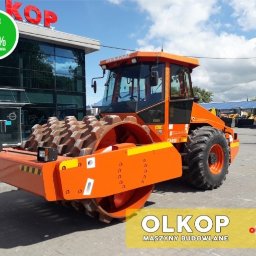 Olkop - Pomarańczowy walec drogowy Dynapac CA612 na placu firmy OLKOP, z widocznym logo firmy i napisem '100% maszyna sprawna' w lewym górnym rogu. W tle budynek firmy z dużymi oknami i inne maszyny...
