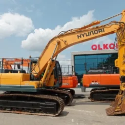 Żółta koparka Hyundai 300LC-9A na placu, widoczne inne maszyny budowlane i budynek z napisem OLKOS w tle.