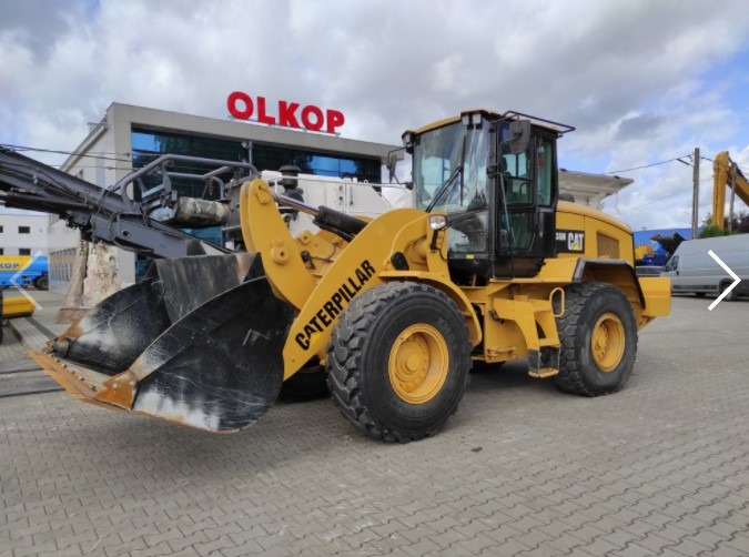 Żółta ładowarka kołowa Caterpillar 330D, zaparkowana na kostce brukowej przed budynkiem z napisem OLKOP, z fragmentem frezarki do asfaltu zamontowanej z przodu.