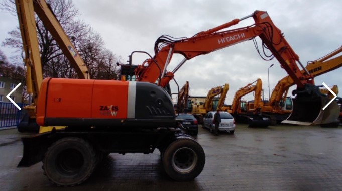 Pomarańczowo-szara koparka kołowa Hitachi Zaxis 140W na placu z innymi maszynami budowlanymi, widoczne samochody osobowe