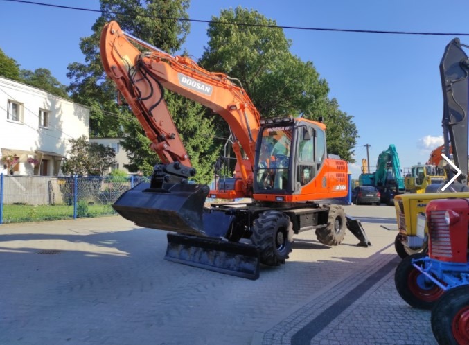 Pomarańczowa koparka kołowa Doosan, zaparkowana w Kowalewie Pomorskim, z widocznymi innymi maszynami budowlanymi w tle.