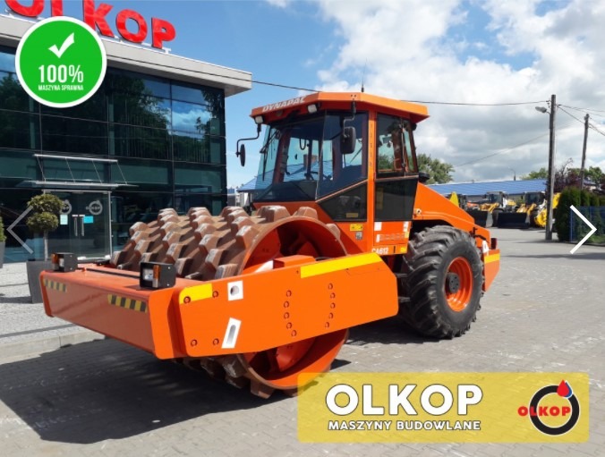 Pomarańczowy walec drogowy Dynapac CA612 na placu firmy OLKOP, z widocznym logo firmy i napisem '100% maszyna sprawna' w lewym górnym rogu. W tle budynek firmy z dużymi oknami i inne maszyny...