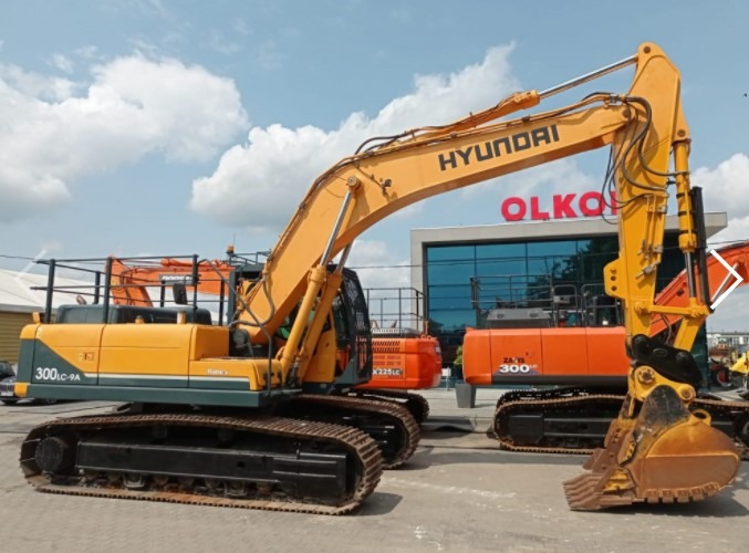 Żółta koparka Hyundai 300LC-9A na placu, widoczne inne maszyny budowlane i budynek z napisem OLKOS w tle.