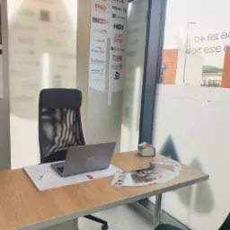 Biuro agenta ubezpieczeniowego: biurko z laptopem, ulotkami i małym modelem domu. Na ścianie logo różnych towarzystw ubezpieczeniowych.