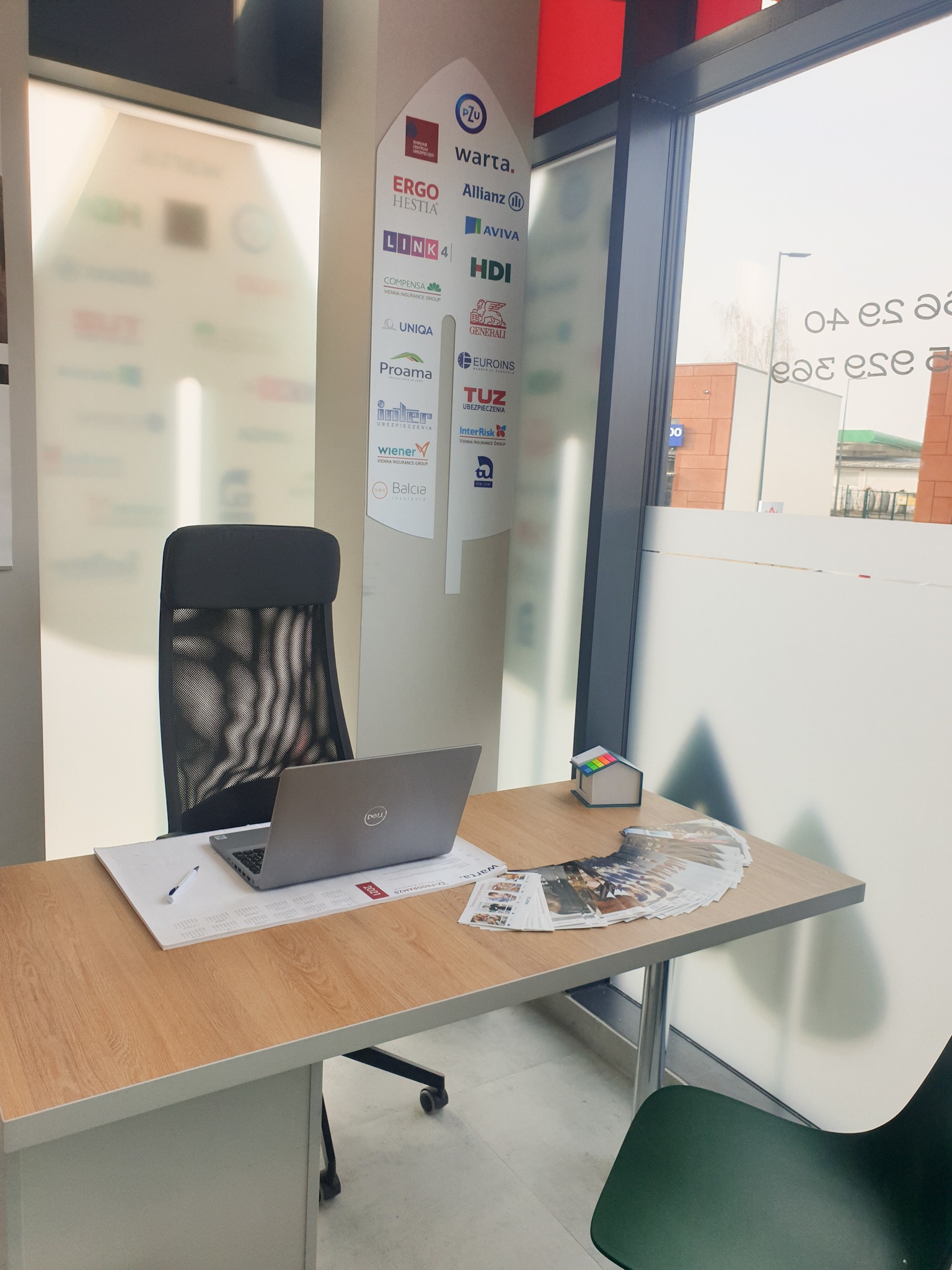 Biuro agenta ubezpieczeniowego: biurko z laptopem, ulotkami i małym modelem domu. Na ścianie logo różnych towarzystw ubezpieczeniowych.