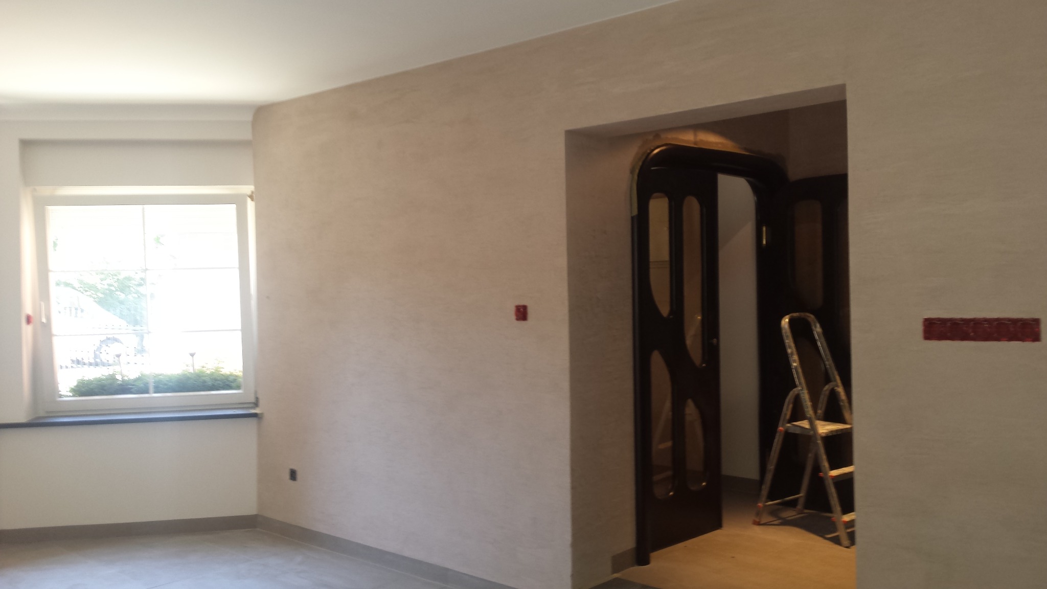 Wnętrze pokoju po nałożeniu tynku dekoracyjnego na ściany, widoczna drabina oparta o ścianę przy otwartych, ciemnych drzwiach.