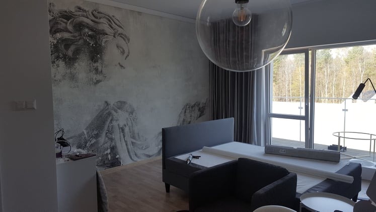 Ściana z szarą tapetą przedstawiającą postać w stylu antycznym w pokoju z szarymi meblami i dużym oknem z widokiem na las.