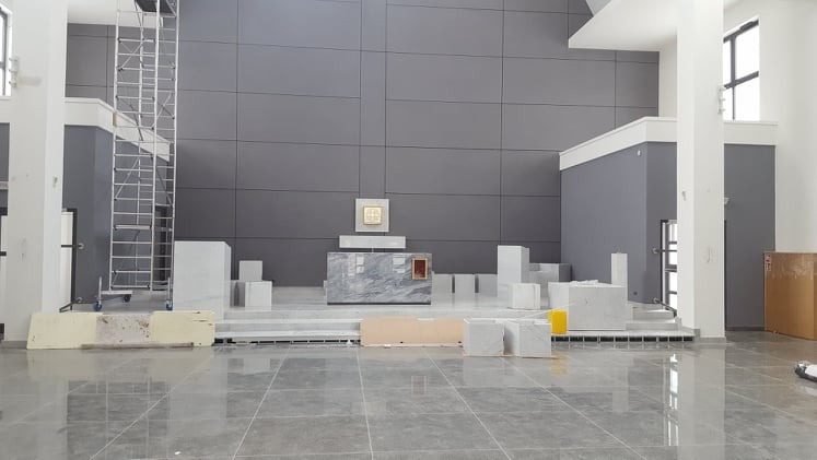 Wnętrze kościoła w trakcie wykańczania, widoczna marmurowa konstrukcja ołtarza, szare panele ścienne, rusztowanie i białe elementy dekoracyjne, posadzka z połyskiem.