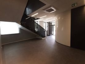 Nowoczesne wnętrze biurowe z widoczną klatką schodową o czarnej konstrukcji i szklanych balustradach, jasnoszara podłoga, białe ściany z elementami dekoracyjnymi w kształcie walca, sufit...