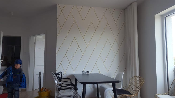 Biała ściana z geometrycznym wzorem w odcieniach złota, czarny stół z krzesłami w jadalni, widok na dziecko w drzwiach.