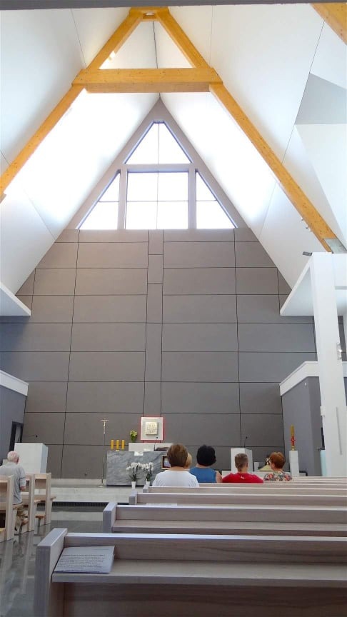 Wnętrze nowoczesnego kościoła z wysoką ścianą wyłożoną szarymi panelami, drewnianą konstrukcją dachu w kształcie trójkąta i rzędami ławek z wiernymi.