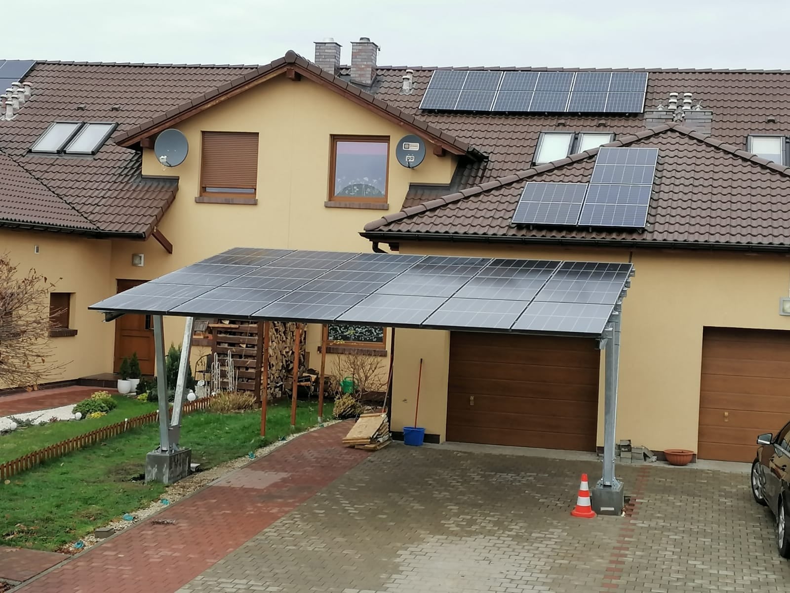 Instalacja paneli słonecznych na dachu domu i jako carport nad wjazdem do garażu, widoczne panele na dachu oraz konstrukcja carportu z szarego metalu, z boku zaparkowany samochód.