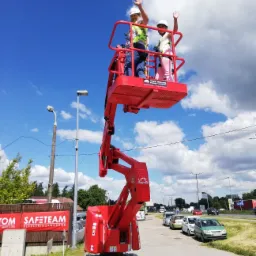 Instruktor i kursantka w kasku i kamizelce na podnośniku koszowym podczas zajęć praktycznych, w tle błękitne niebo z chmurami i budynek z banerem reklamowym