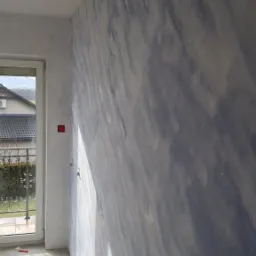 Ściana w pokoju po nałożeniu dekoracyjnej gładzi w odcieniach bieli i błękitu, z widocznym oknem i fragmentem podłogi w surowym stanie.