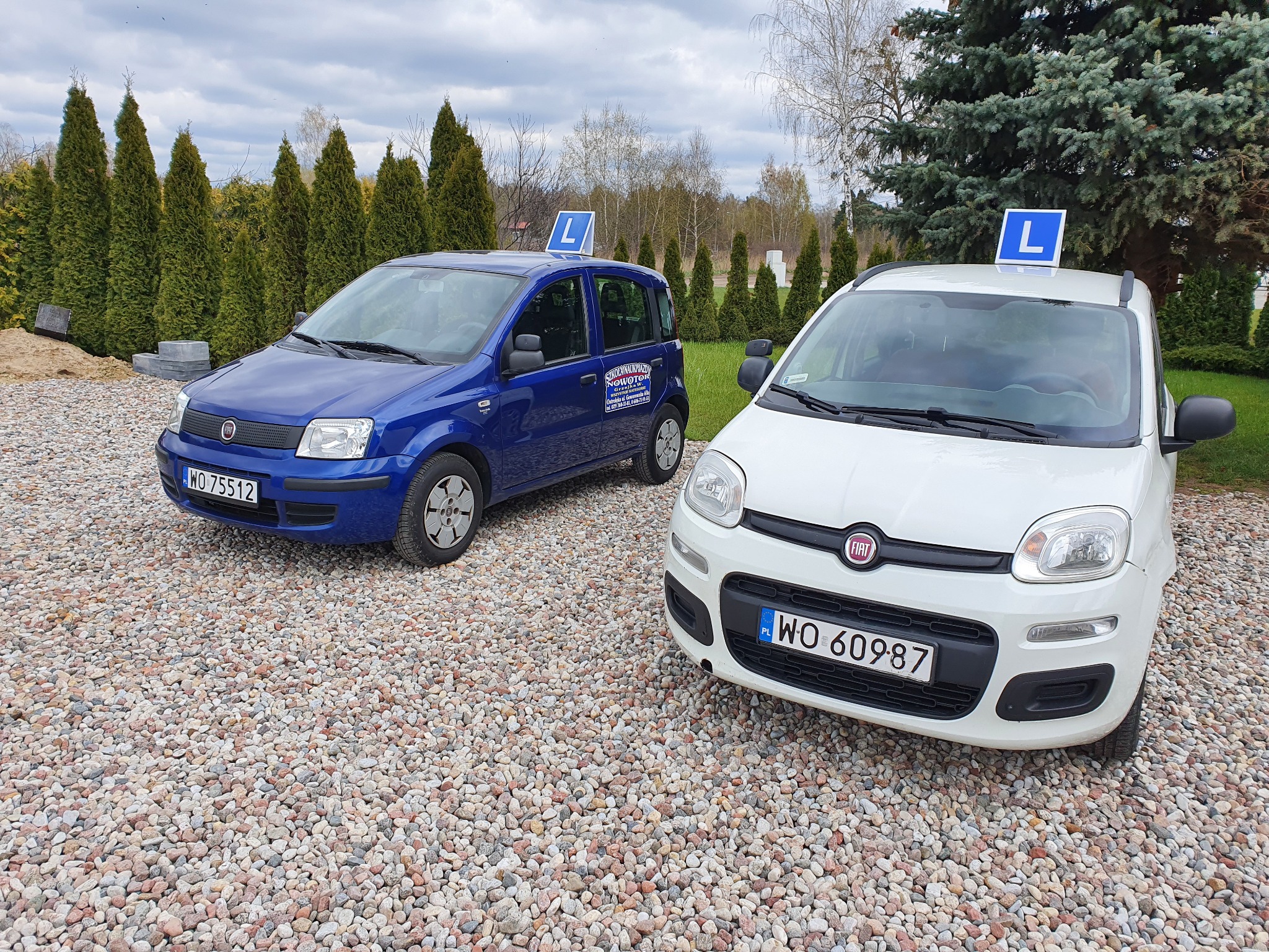 Dwa samochody Fiat Panda, niebieski i biały, z oznaczeniem 'L' na dachu, stojące na żwirowym podłożu przed zielonymi tujami, używane w szkole jazdy.