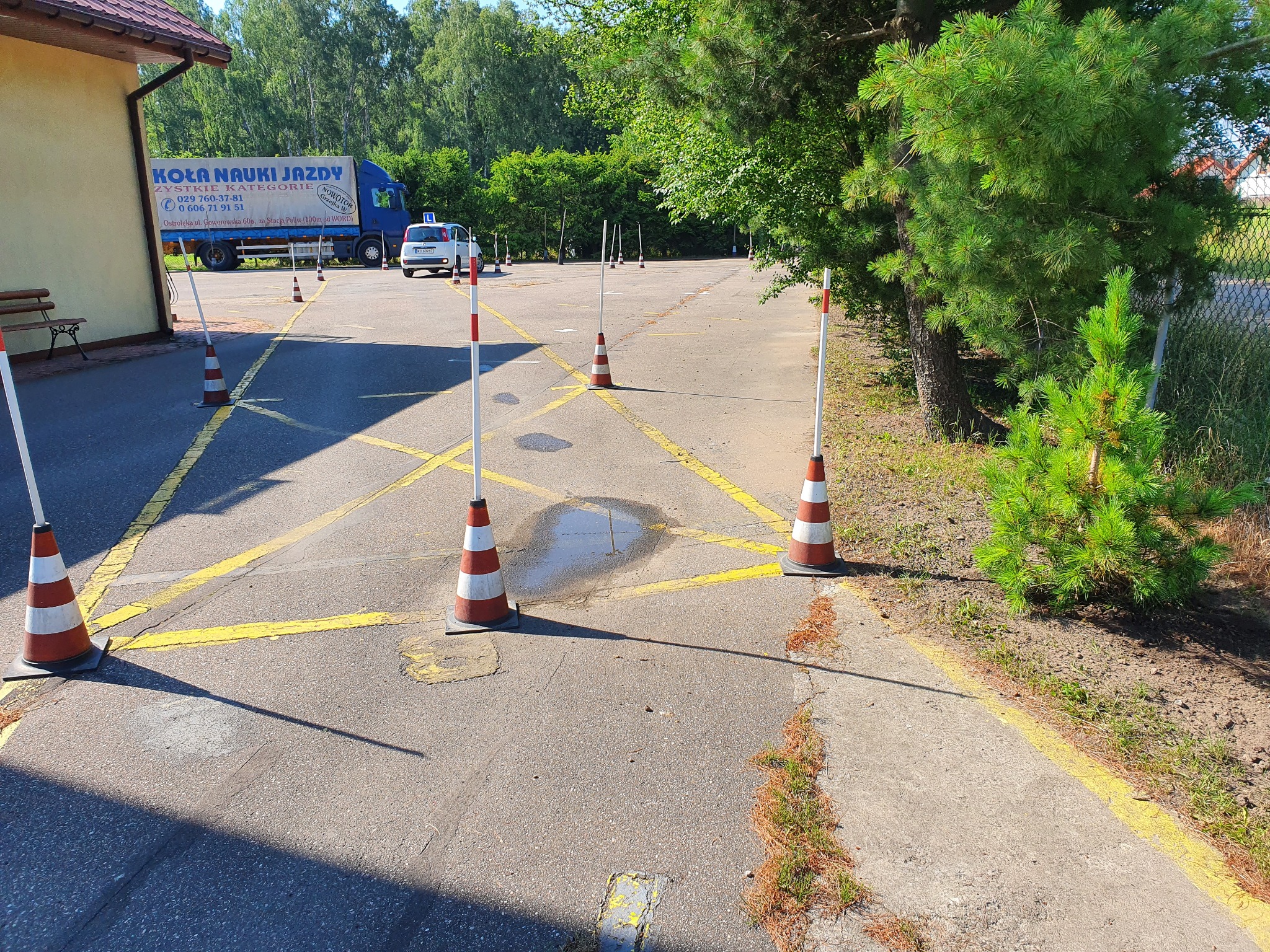 Plac manewrowy z wyznaczonymi żółtymi liniami i ustawionymi pachołkami, w tle budynek, ciężarówka z reklamą i samochód z literą 'L'.