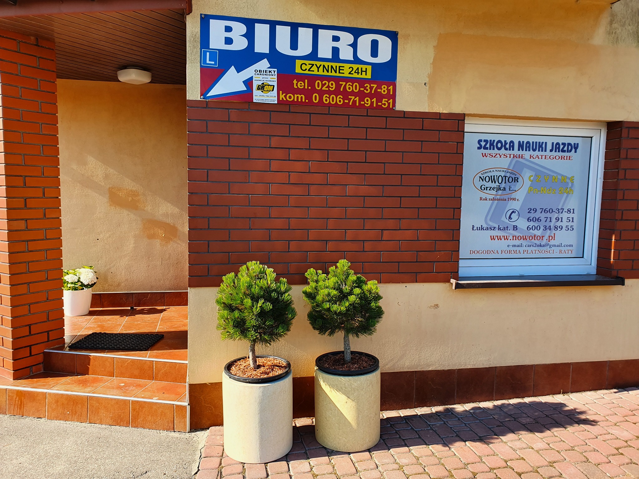 Wejście do biura z szyldem 'BIURO' i reklamą 'Szkoła Nauki Jazdy NOWOTOR', dwie małe tuje w donicach przed budynkiem.