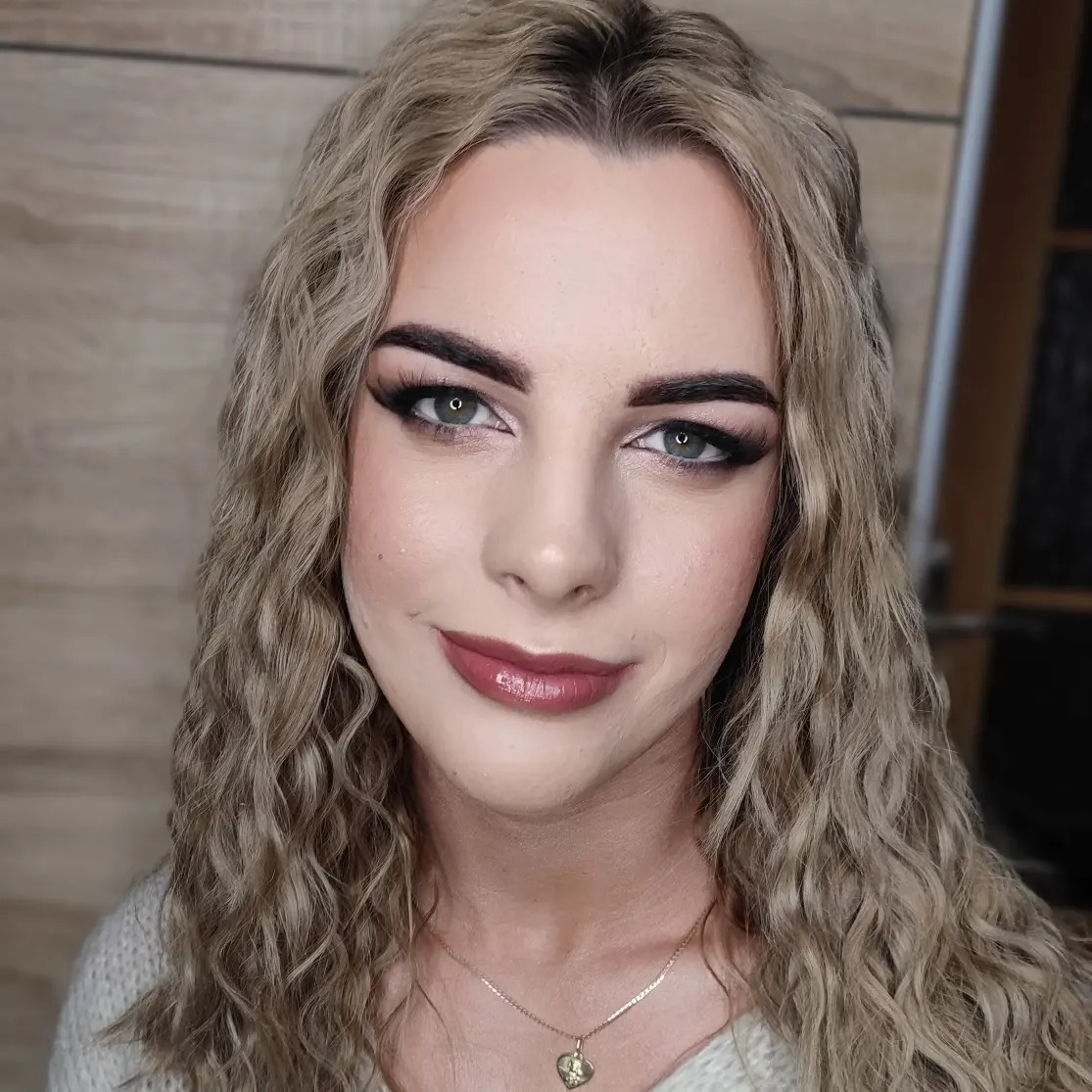 Portret kobiety z profesjonalnym makijażem: smoky eye z długimi rzęsami, błyszczące usta w odcieniu burgundu, podkreślone brwi i delikatny kontur twarzy. Długie, blond, kręcone włosy opadają...