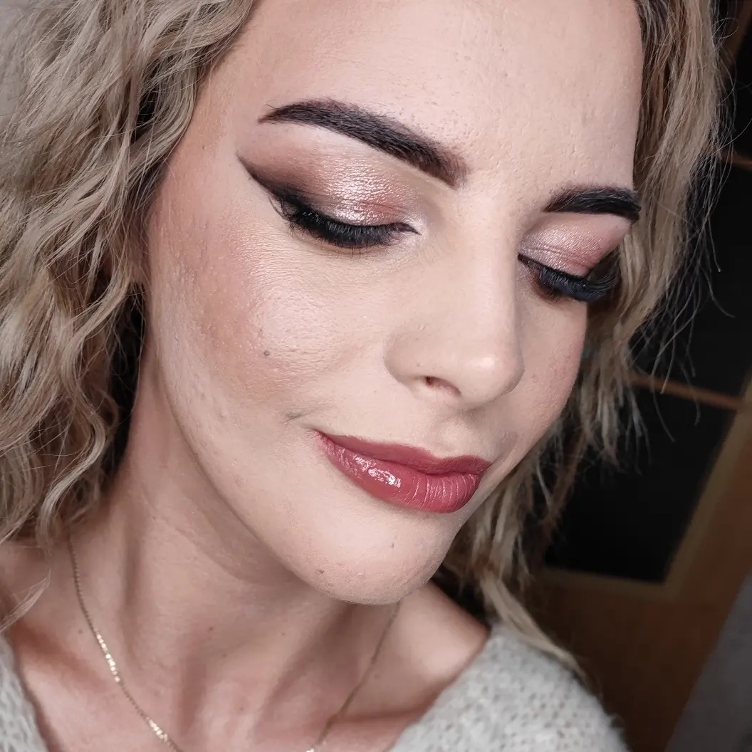 Kobieta z zamkniętymi oczami prezentuje delikatny, różowo-złoty cień do powiek z wyraźną czarną kreską, podkreślone brwi i błyszczące, bordowe usta. Widoczny delikatny makijaż policzków i złoty...