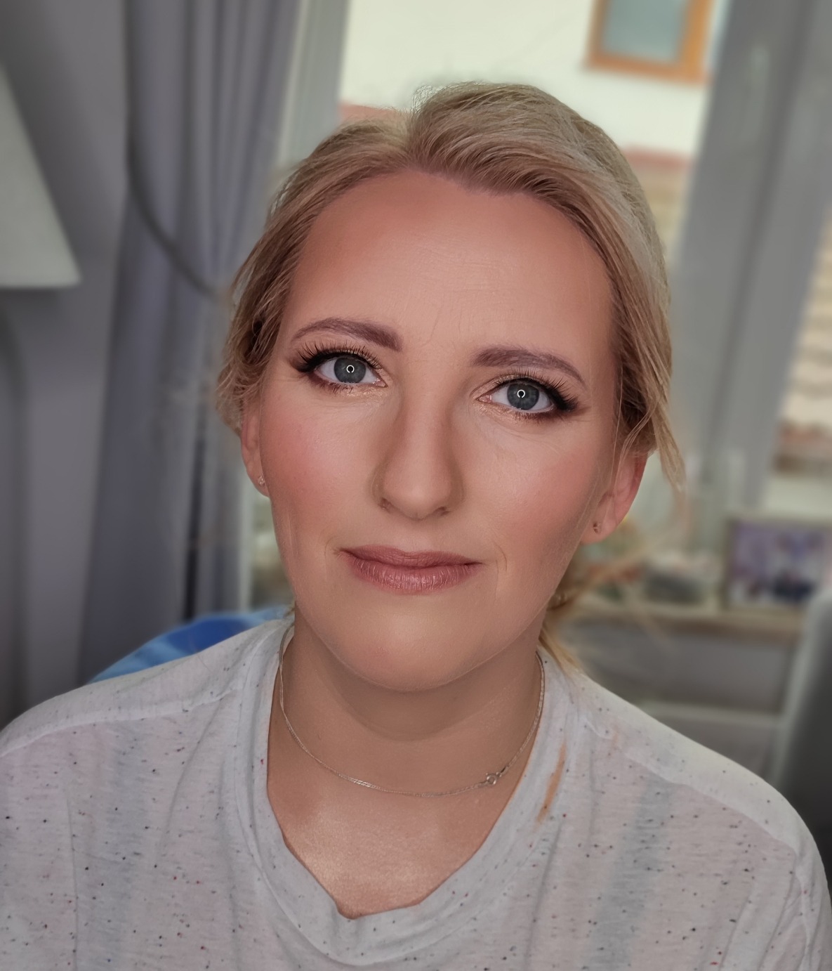 Portret kobiety z delikatnym, dziennym makijażem, z podkreślonymi oczami i różem na policzkach, w białej koszulce z drobnymi plamkami, srebrny łańcuszek na szyi.