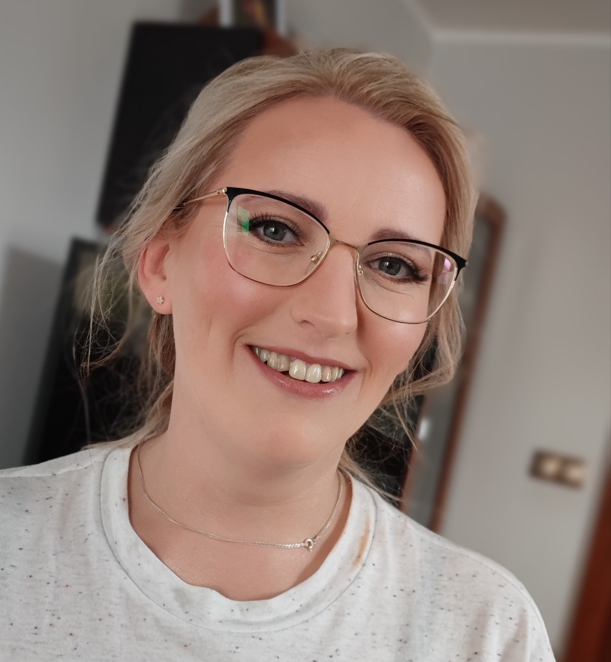 Uśmiechnięta blondynka w okularach z delikatnym makijażem dziennym, podkreślone brwi i róż na policzkach, widoczna srebrna celebrytka na szyi i mała kolczyk-gwiazdka w uchu.