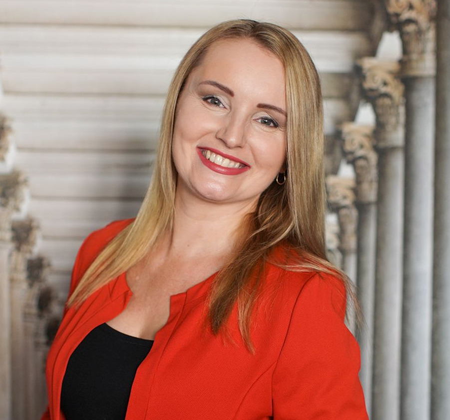 Uśmiechnięta blond kobieta w czerwonej marynarce na tle kolumn, prezentująca pewność siebie i profesjonalizm.