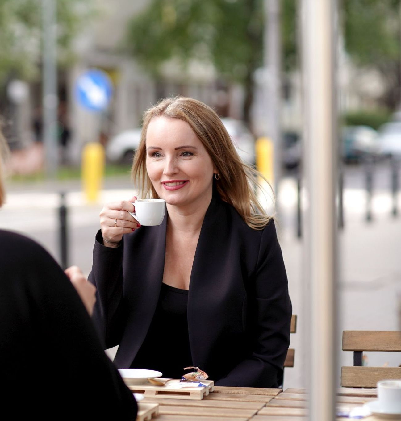 Kobieta w czarnym żakiecie, siedząca przy drewnianym stole na zewnątrz, pije kawę z białego kubka i uśmiecha się, w tle widoczny ruch uliczny i znak drogowy.