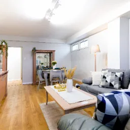 Jasne wnętrze salonu w Łodzi z drewnianą podłogą, szarą sofą z dekoracyjnymi poduszkami, stolikiem kawowym z wazonem i komodą z lustrem w tle.