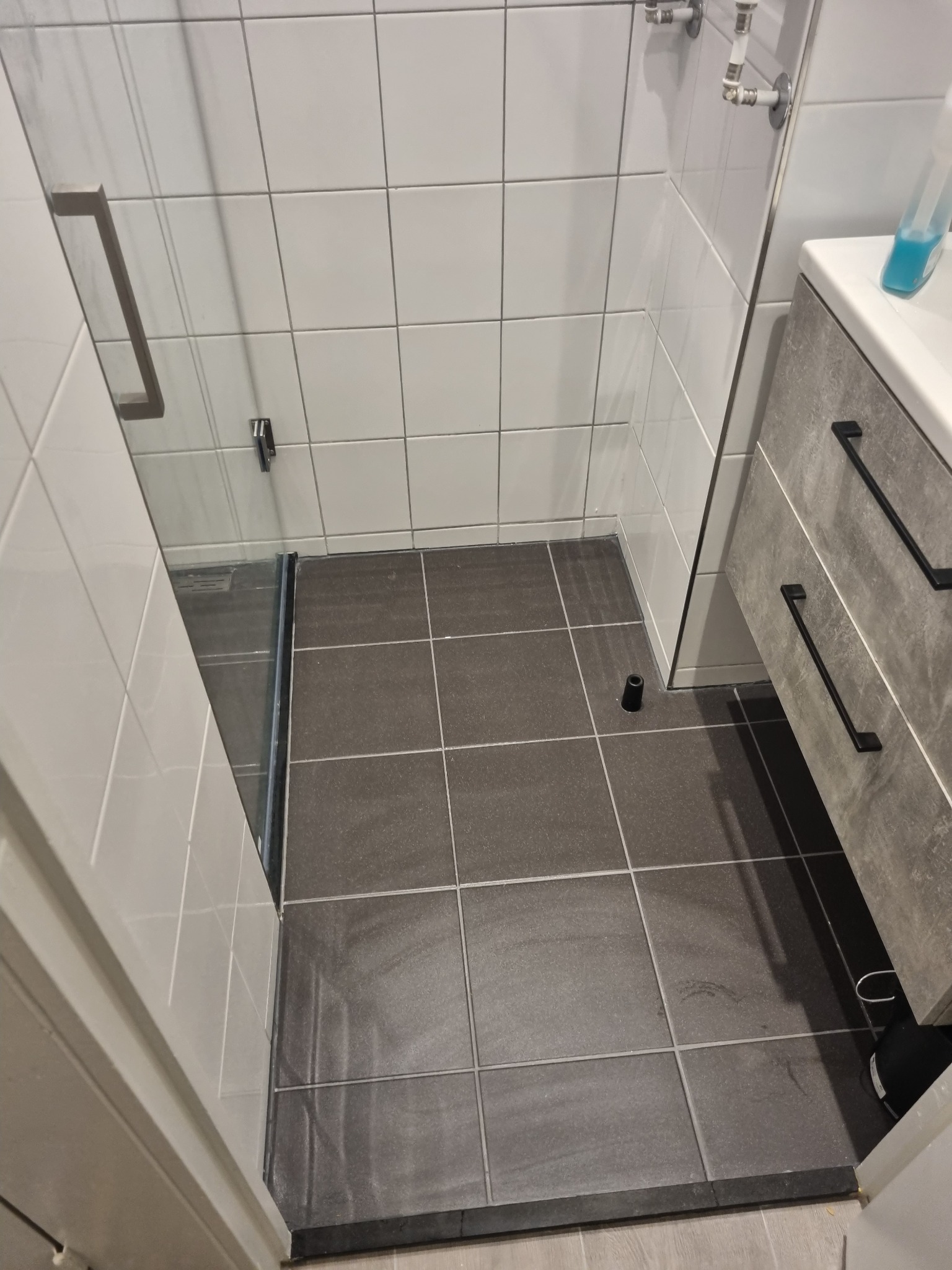 Nowoczesna kabina prysznicowa z szarymi płytkami podłogowymi i białymi płytkami ściennymi, widoczna szklana ściana kabiny oraz umywalka z szufladami w szarym kolorze.