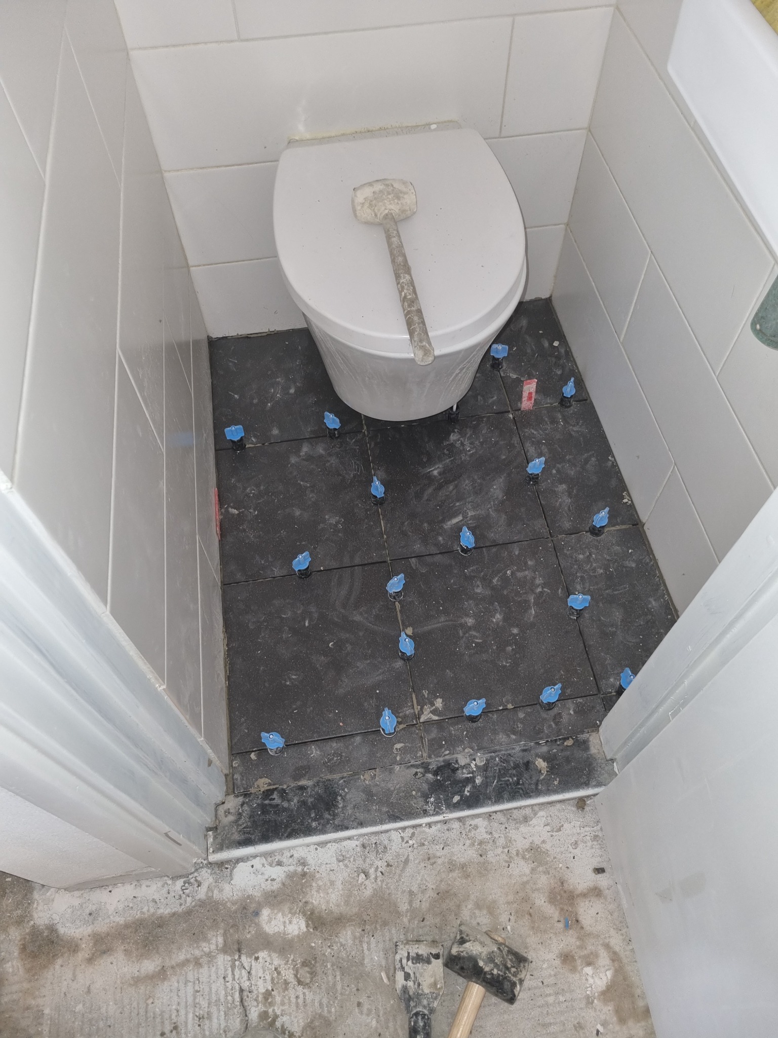 Układanie ciemnych płytek podłogowych w małej łazience, widoczne niebieskie klipsy poziomujące, szpachelka na sedesie i młotek na podłodze.