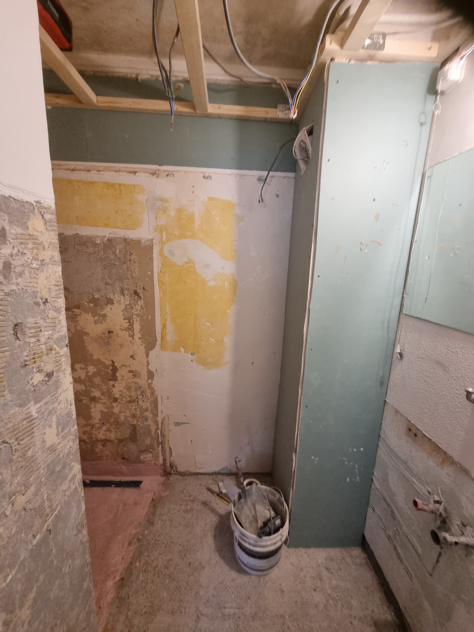 Wnętrze łazienki w trakcie prac remontowych, widoczne odsłonięte ściany z pozostałościami starej farby i tynku, instalacja elektryczna pod sufitem, płyta gipsowo-kartonowa oparta o ścianę...