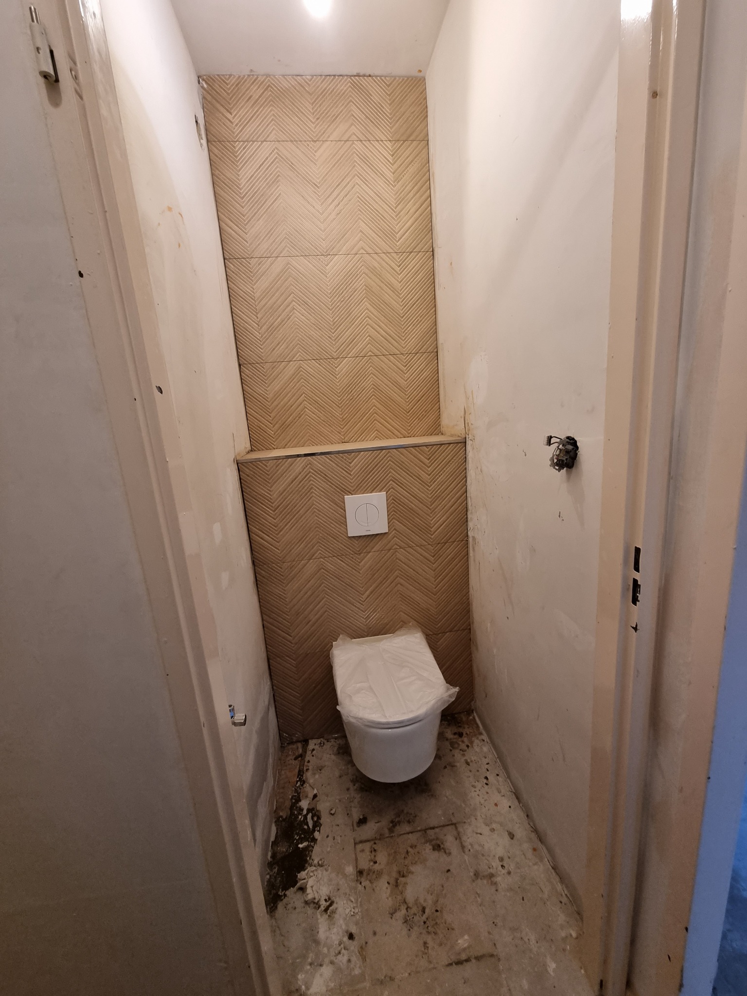 Remont toalety w trakcie: położone płytki chevron na ścianie, zamontowana podwieszana miska WC z przykryciem folią, ściany w trakcie szpachlowania, widoczne niedoskonałości podłogi.