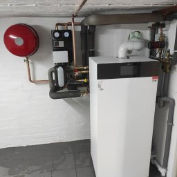 Nat-Bud - Nowoczesny piec gazowy Viessmann z podłączoną instalacją grzewczą, widoczne rury miedziane, manometry i zawory, zamontowany w pomieszczeniu z szarą podłogą i białymi ścianami.