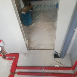 Nat-Bud - Czerwone rury instalacji grzewczej z miedzianymi złączkami, zamontowane na surowej ścianie w trakcie prac wykończeniowych, widok z góry.