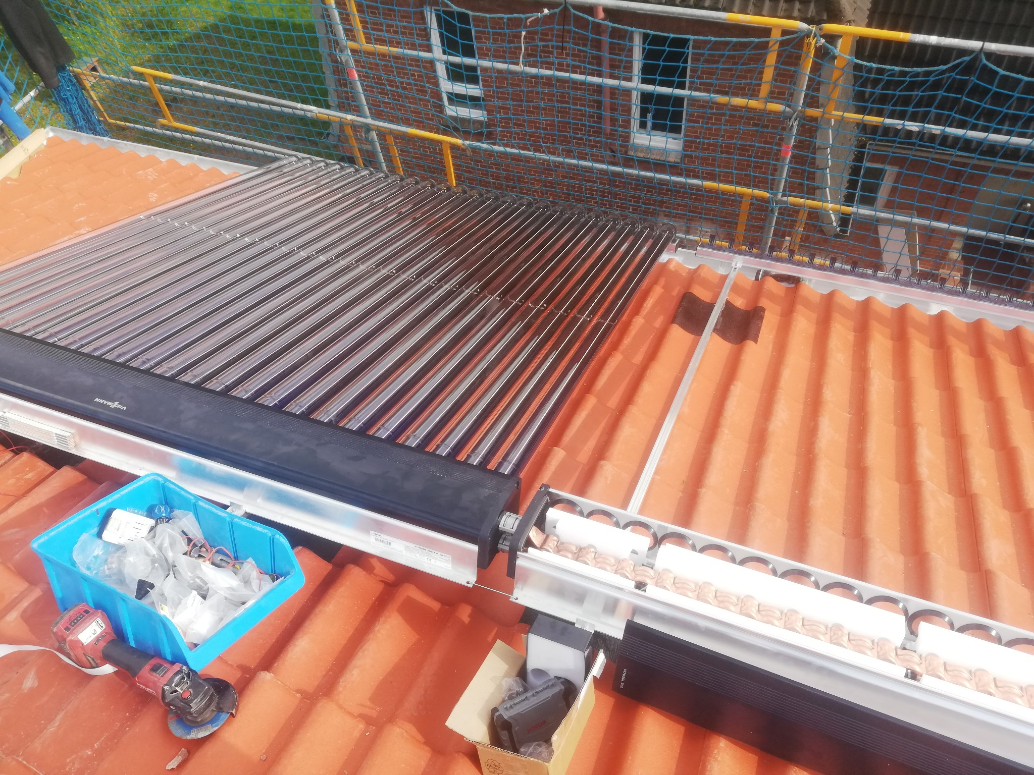 Montaż kolektorów słonecznych na dachu z czerwonej dachówki, widoczne narzędzia i akcesoria w niebieskim pojemniku, zabezpieczenia rusztowania w tle.