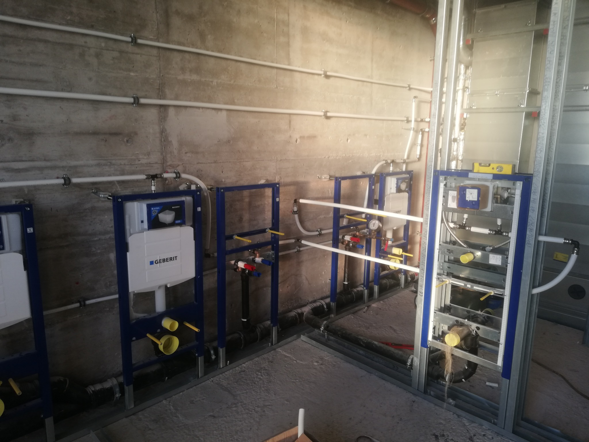 Surowy beton i widok na instalację podtynkową WC z niebieskimi ramami, rurami doprowadzającymi wodę i odpływami kanalizacyjnymi w trakcie montażu.