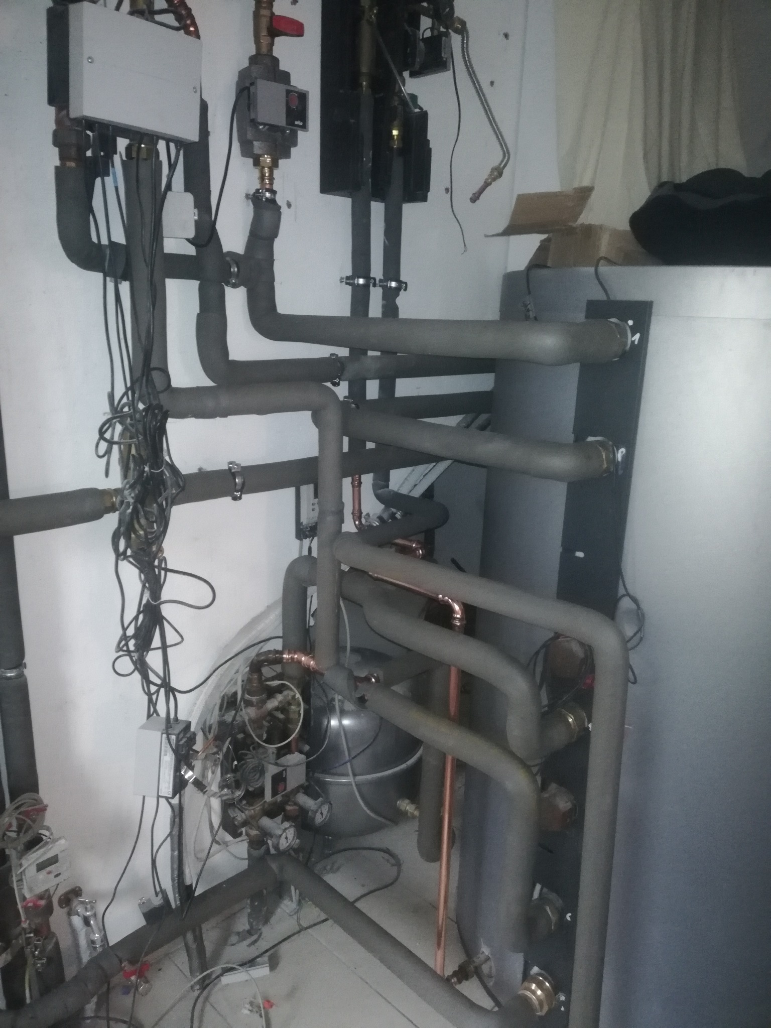 Skrzyżowanie rur instalacji centralnego ogrzewania z widocznymi zaworami, manometrami i przewodami elektrycznymi w pomieszczeniu technicznym.