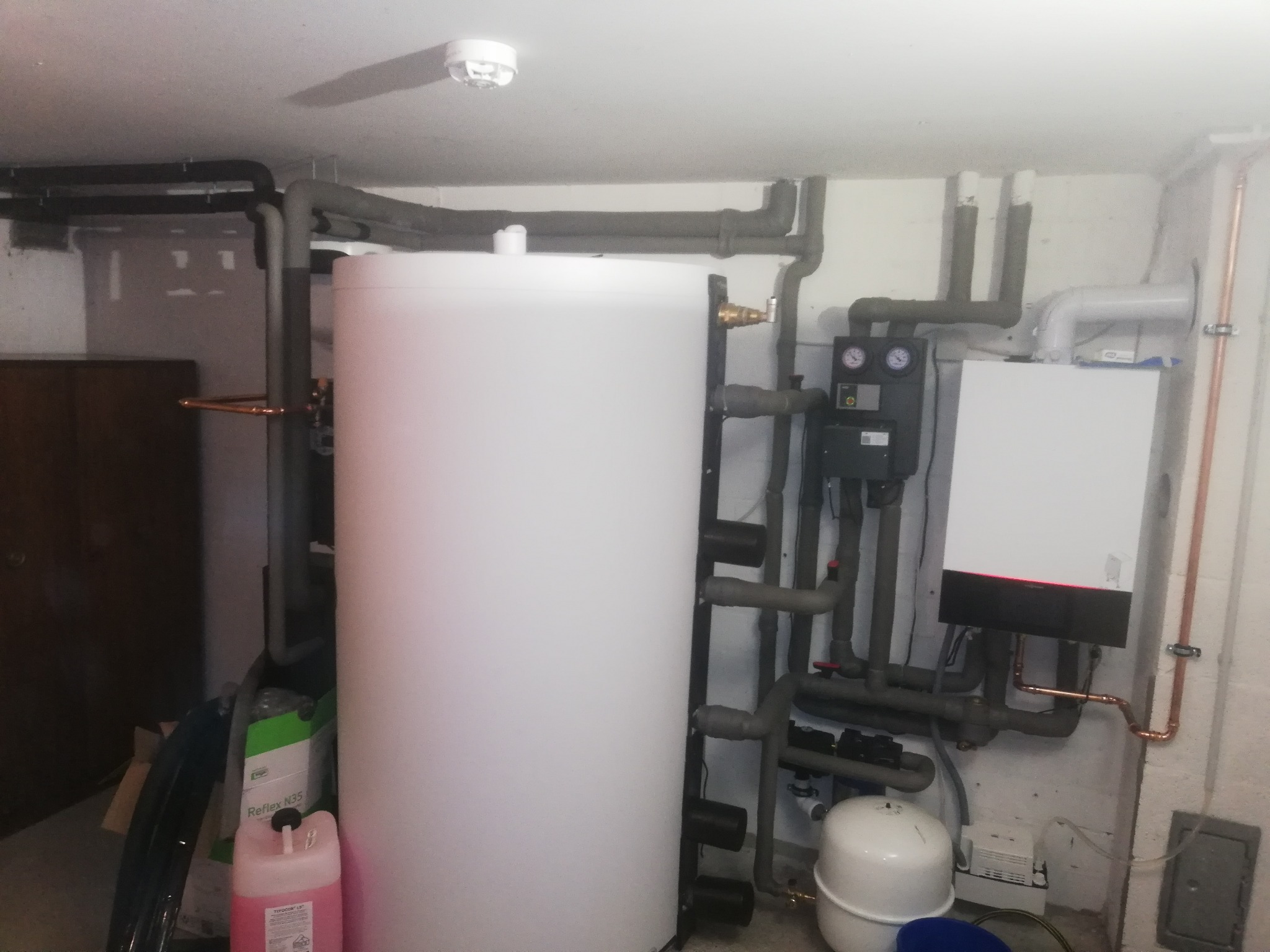 Biały zasobnik ciepłej wody z podłączoną instalacją grzewczą, widoczne rury miedziane i izolowane szare rury, ekspansomat, sterownik i piec gazowy w kotłowni. Na podłodze płyn do instalacji...
